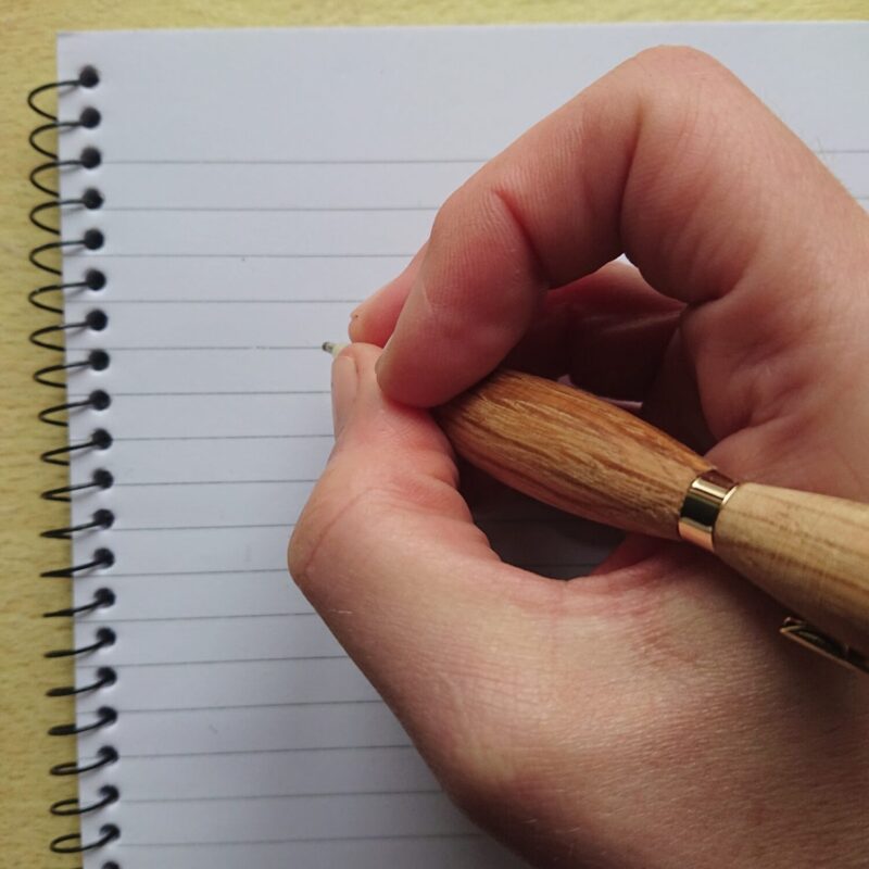 oak handmade pen with wider grip, but narrow waist being held to write. The pen has a retractable tip.